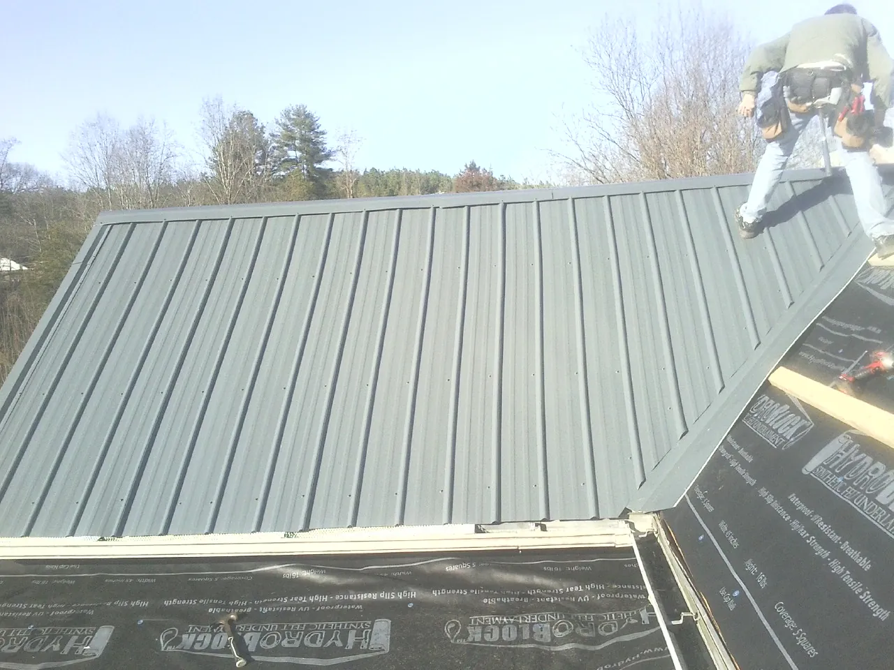 Crew installing dark green standing seam panels over synthetic underlayment in Fair Haven
