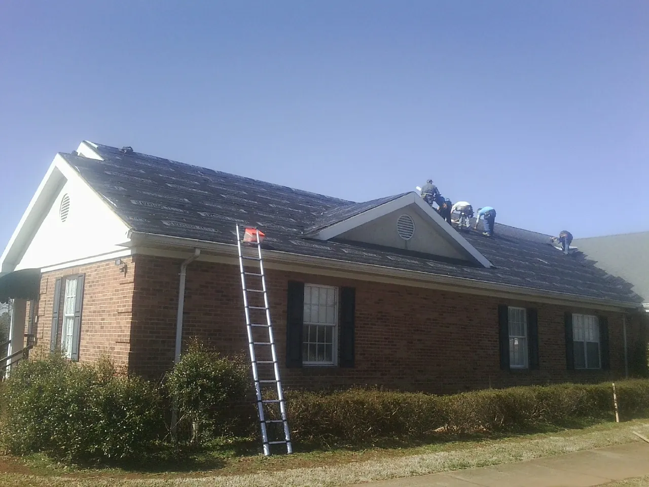 Crew tearing shingles off a brick ranch roof with a ladder set — Metal Roof Repair work by our crew in Fair Haven