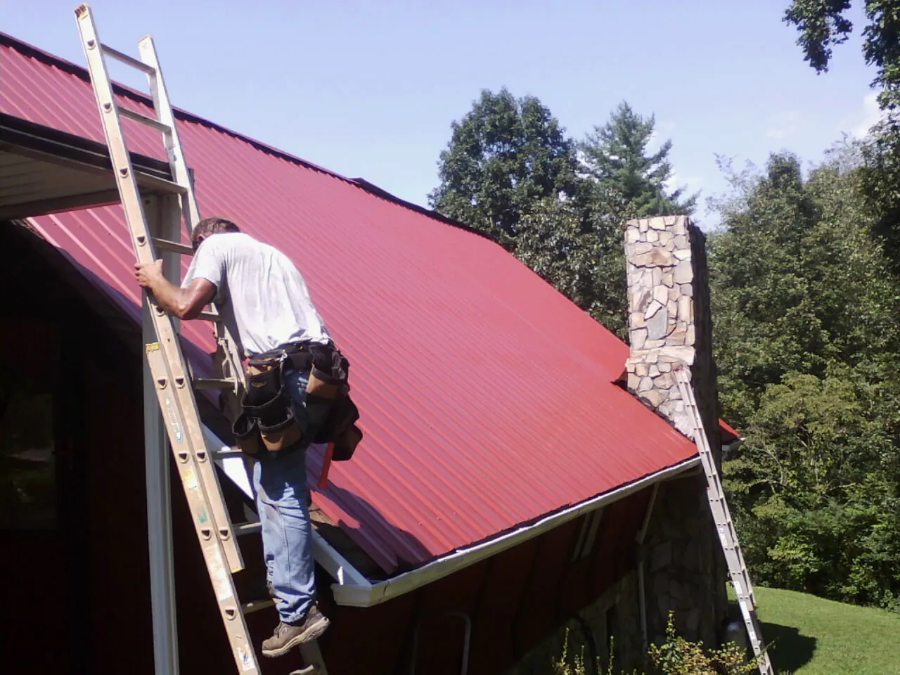 Roofer on a ladder beside a red metal roof and stone chimney — Soffit & Fascia Repair by Roofing Brothers in Fair Haven